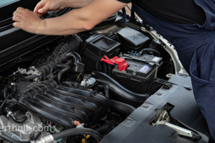 Gari ke engine compartment ka scene, jahan aik mechanic leisure battery ya main battery ko check kar raha hai. Red terminal visible hai.