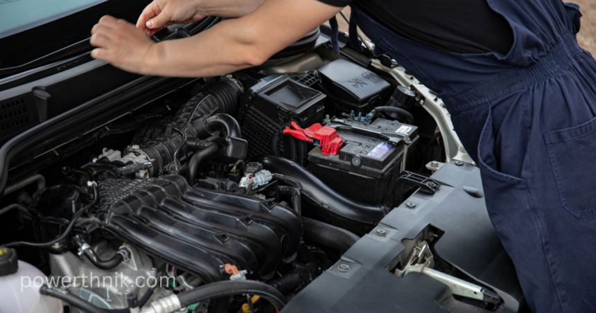 Gari ke engine compartment ka scene, jahan aik mechanic leisure battery ya main battery ko check kar raha hai. Red terminal visible hai.