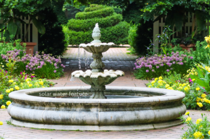 Solar water feature three-tier stone fountain in a circular basin surrounded by yellow marigolds and purple alliums in a brick-paved garden.